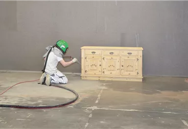 A person in protective gear is working on a wooden cabinet. The cabinet is on uneven ground in an open space.