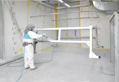 A worker in protective clothing is spraying paint on a metallic frame in a clean, well-ventilated room. The floor is light and the walls are gray with yellow markings.
