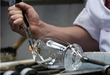 A craftsman is shaping a glass with a pair of tongs. In the background, tools for glass processing are visible.