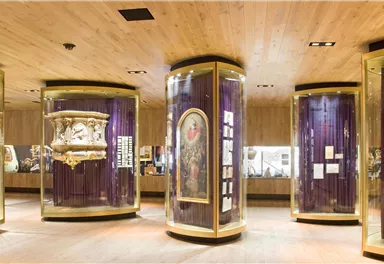 A modern exhibition space with display cases showcasing various art historical objects. The walls are in warm wood tones, and the lighting highlights the exhibits.