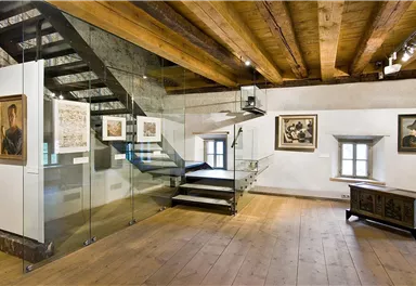 A modern exhibition space with a glass staircase and wooden ceilings. Several framed pictures hang on the walls.