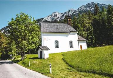 A small, white church stands surrounded by green meadows and trees. In the background, majestic mountains can be seen under a clear sky.
