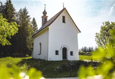A small, white church surrounded by greenery and trees. The sky is blue with a few clouds.