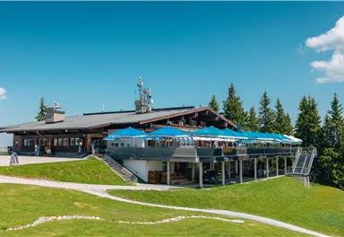 A modern restaurant in the mountains with a large terrace and blue sun umbrellas. Surrounded by green meadows and coniferous trees under a clear blue sky.
