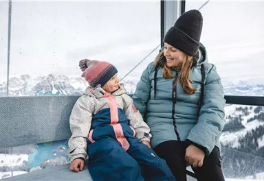 Eine Mutter und ihr Kind sitzen in einer Gondel mit Blick auf schneebedeckte Berge. Beide tragen warme Winterkleidung und lächeln sich an.