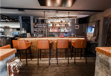 A cozy bar with a wooden counter and modern bar stools. In the background, there are shelves with drinks and warm lighting.