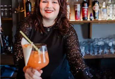 A friendly bartender smiles and holds two colorful cocktails in her hand. In the background, shelves filled with various spirits are visible.