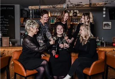 A group of six women is celebrating in a bar. They are toasting with glasses and are dressed cheerfully.