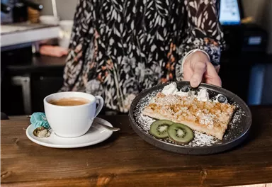 A person is handing a piece of cake with fruit on a plate. Next to it, there is a cup of coffee on the table.