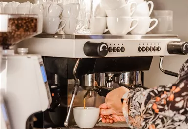 A person is preparing coffee at an espresso machine in a café. In the background, numerous cups and glasses are visible.