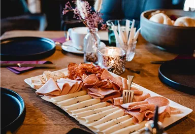 A beautifully set table with various types of sausages and cheeses on a platter. In the background, cozy seating and decorations are visible.