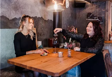 Two women are sitting at a wooden tray in a modern café. One woman is pouring something into a glass while the other is watching kindly.
