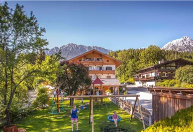 Ein malerisches Bergdorf mit zwei gemütlichen Holzhäusern. Kinder spielen auf einem Spielplatz im Grünen, umgeben von schöner Natur.