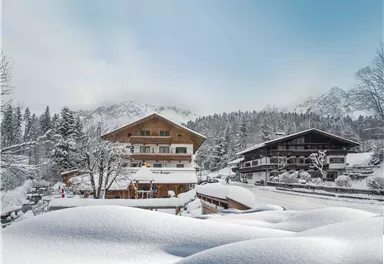 Eine verschneite Landschaft mit traditionellen Holzchalets. Im Hintergrund sind schneebedeckte Berge und ein klarer Himmel zu sehen.