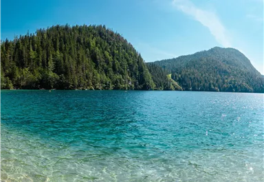 A clear blue lake, surrounded by green forests and mountains. The sun is shining, creating a peaceful atmosphere.