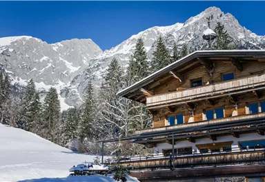 A charming chalet in the snow, surrounded by tall mountains and coniferous trees. The sky is clear and blue, creating a wintry atmosphere.