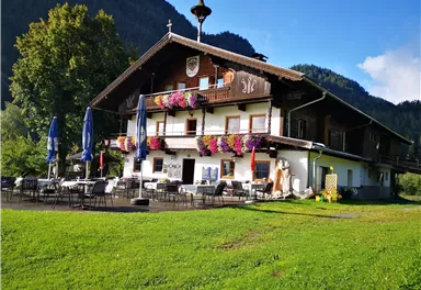 Ein traditionelles Holzhaus mit bunten Blumen an den Fenstern. Es liegt in einer grünen Umgebung mit Bergen im Hintergrund.