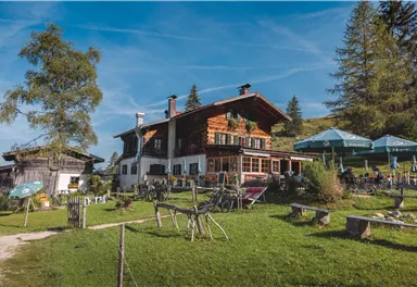 A rustic mountain house surrounded by a green meadow and trees. In the foreground, seating areas and sun umbrellas can be seen.