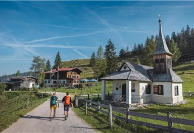 Two hikers are walking on a gravel path in a picturesque landscape. In the background, a small chapel and traditional mountain houses can be seen.