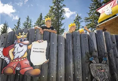 Two children with crowns are standing by a wooden fence. Next to them is a colorful sign featuring a royal figure and the inscription "KaiserWelt".
