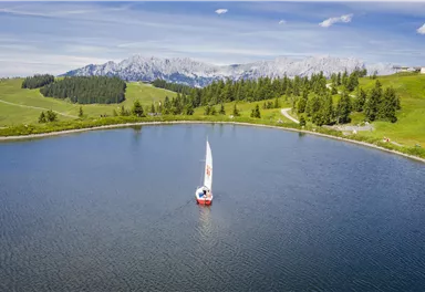 A picturesque lake surrounded by gentle hills and trees. A sailboat glides smoothly over the water while the mountains are visible in the background.