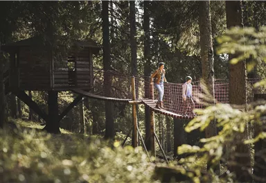 A woman and a child are walking on a suspension bridge between trees. In the background, a treehouse can be seen.