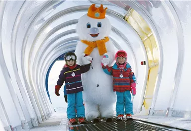 Zwei Kinder stehen neben einem großen Schneemann in einem winterlichen Gang. Sie tragen bunte Winterkleidung und lächeln fröhlich.