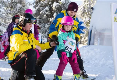 Ein fröhliches Kind lernt mit Hilfe eines Trainers das Skifahren. Es steht an der Startlinie in einer verschneiten Landschaft.