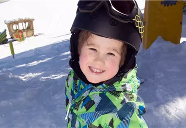 Ein lächelndes Kind im Schnee trägt einen Helm und eine bunte Jacke. Im Hintergrund sind Spielgeräte und die schneebedeckte Landschaft zu sehen.