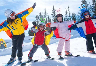 Eine Skilehrerin und drei Kinder stehen auf Skiern im Schnee. Alle tragen bunte Skikleidung und Helme, während sie fröhlich posieren.