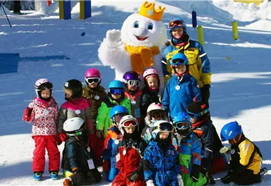 Eine Gruppe von Kindern in bunten Skianzügen steht im Schnee und lächelt. Im Hintergrund ist ein großer, freundlicher Schneemann mit einer Krone zu sehen.