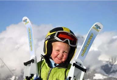 Ein lächelndes Kind trägt einen Helm und hält Skier in den Händen. Im Hintergrund sind schneebedeckte Berge und ein blauer Himmel.