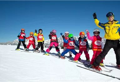 Eine Gruppe von Kindern steht auf Skiern im Schnee und lächelt. Ein Skilehrer winkt ihnen daneben zu.