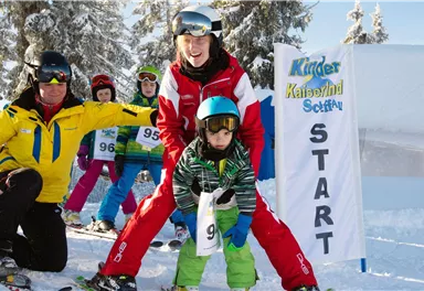 Ein fröhliches Skirennen für Kinder mit zwei Lehrern und mehreren kleinen Skifahrern. Im Hintergrund ist das Startbanner gut sichtbar.