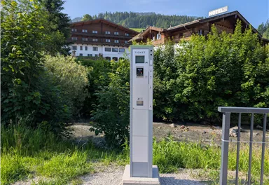 Ein Parkautomaten steht neben einem Fluss, umgeben von Bäumen. Im Hintergrund sind Holzgebäude und ein Hotel sichtbar.