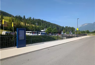 Eine ruhige Straße mit einer grünen Hügelkulisse und Bäumen. Auf der linken Seite steht ein Infostand.