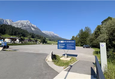 Ein Parkplatz in den Bergen mit klarer Sicht auf die umliegenden Gipfel. Der Himmel ist blau und die Umgebung ist grün und einladend.