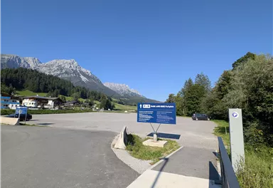 Ein malerischer Parkplatz in den Bergen mit klar blauem Himmel. Im Hintergrund sind hohe Berge und grüne Wälder zu sehen.