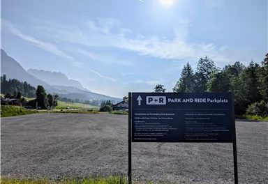 Ein Parkplatz für Park-and-Ride mit einem großen Hinweisschild. Im Hintergrund sind Berge und ein klarer Himmel zu sehen.