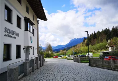 An idyllic view of a well-maintained building with the inscription "Scheffau am Wilden Kaiser". In the background, green mountains rise under a cloudy sky.