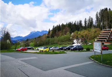 A picturesque landscape with mountains in the background and a parking lot full of cars. The surroundings are green with trees and meadows.