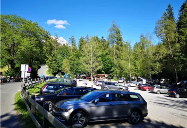 A parking lot surrounded by trees and green countryside. Several cars are parked under a clear blue sky.