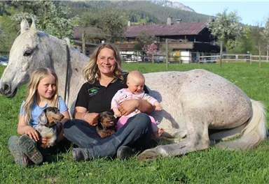 A woman is sitting on a meadow with two children. Next to them lies a white horse and they are holding two small dogs.