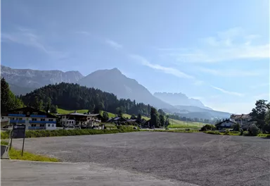 Eine ruhige Landschaft mit Bergen im Hintergrund und einem klaren Himmel. Im Vordergrund sind einige Gebäude und eine große, leere Fläche zu sehen.
