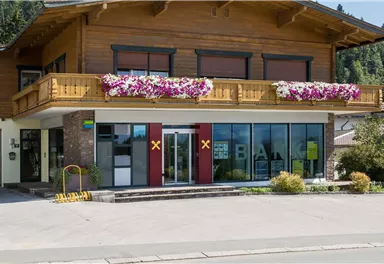 Ein modernes Bankgebäude mit Holzverkleidung und Balkon. Bunte Blumen schmücken die Fassade, umgeben von einem grünen Garten.