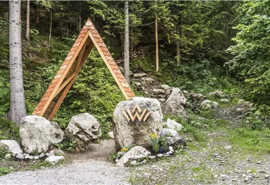 Ein hölzernes Dreiecksschild steht zwischen Bäumen und großen Steinen. Der Weg führt durch eine grüne, bewaldete Landschaft.