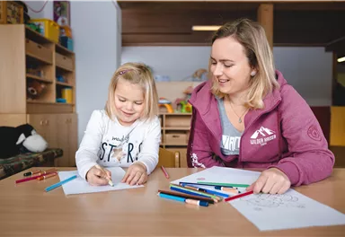 Ein fröhliches Mädchen und eine junge Frau sitzen an einem Tisch und zeichnen. Bunte Stifte und Papiere liegen vor ihnen.