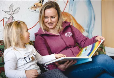 Eine Frau liest einem kleinen Mädchen ein Buch vor. Im Hintergrund sind bunte Wandmalereien zu sehen.