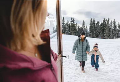 Eine Mutter und ihr Kind kommen fröhlich aus dem Haus in die verschneite Landschaft. Im Hintergrund sind hohe Bäume und ein grauer Himmel zu sehen.
