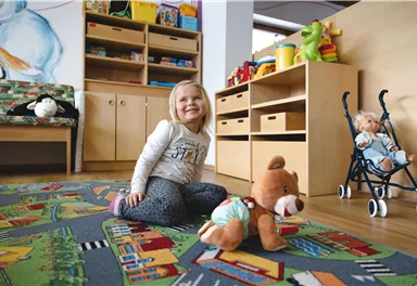 Ein fröhliches kleines Mädchen sitzt auf einem bunten Teppich in einem Kinderzimmer. Um sie herum sind Spielzeuge und Möbel für Kinder.